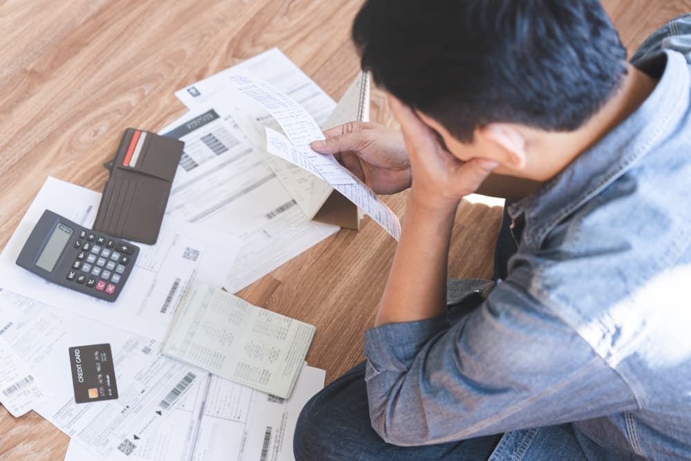 Financial struggle concept: A young Asian man sits stressed and confused, overwhelmed by calculating expenses from credit card bills and invoices, unable to pay his mortgage or loan, facing debt or potential bankruptcy.







